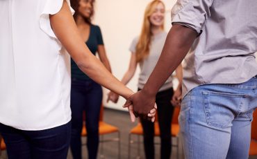 Multi-Cultural Group Of Men And Women Joining Hands At Mental Health Group Therapy Meeting